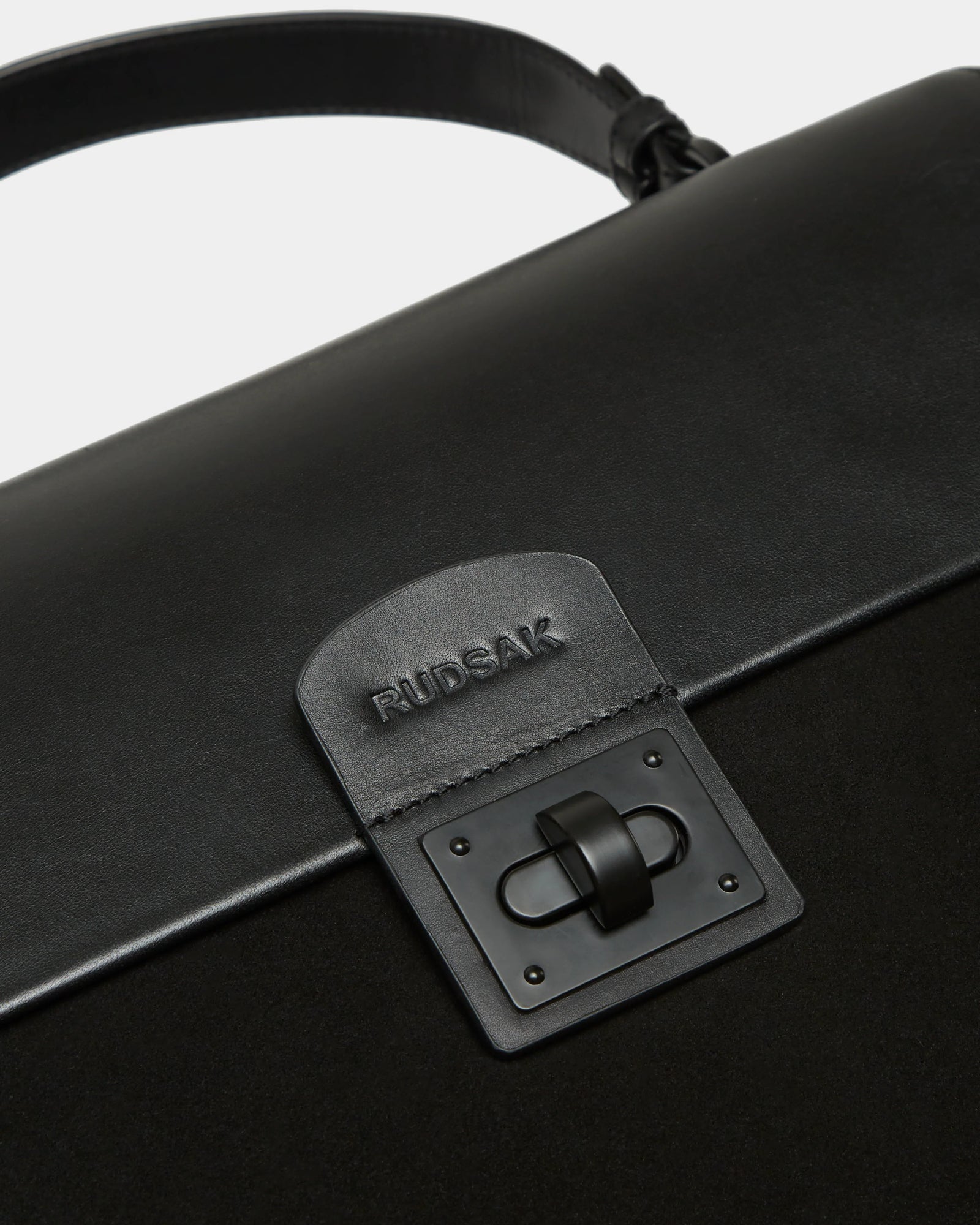 Close-up of the Rudsak ESMA - 8324519 Black handbag, featuring suede and leather, a front flap with metal clasp, and an embossed logo patch above the lock.