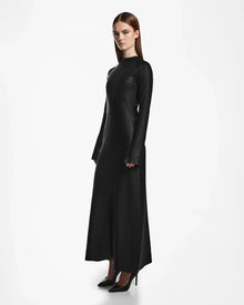 A woman with straight brown hair stands against a white background, wearing the Rudsak FAIZA - 8524579 Black fitted maxi dress with long sleeves and black high heels, facing slightly to the side with a neutral expression.