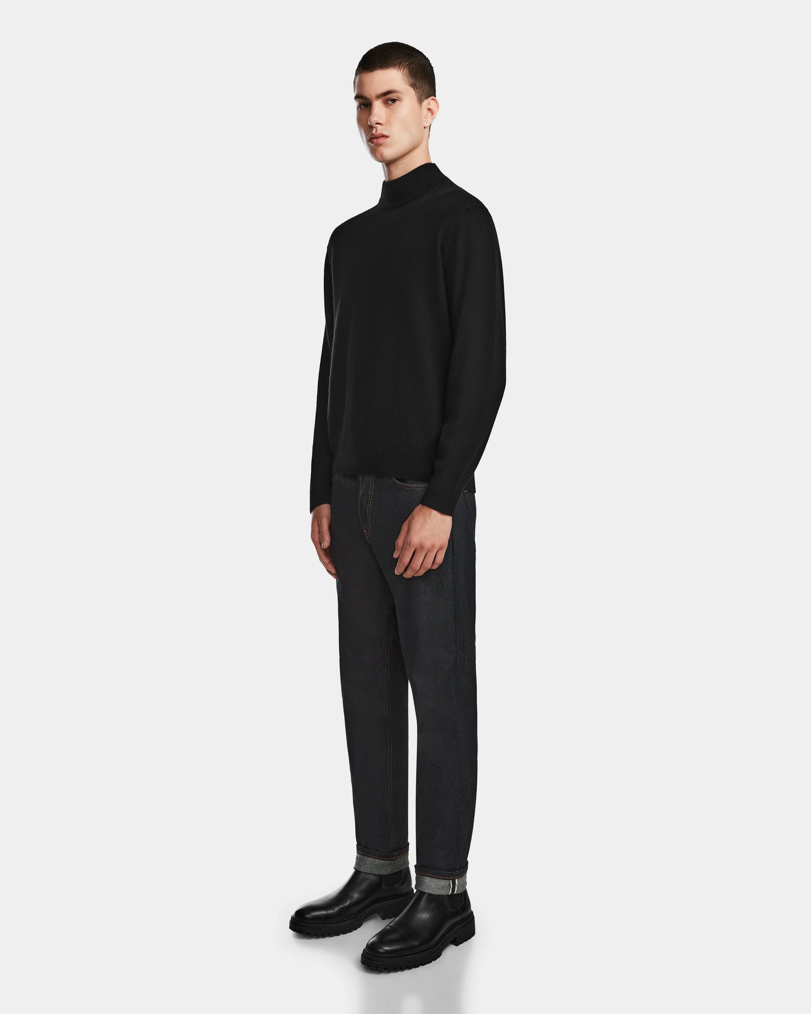 A young man stands against a white background wearing the Rudsak FERRAN - 6524511 Black mens turtleneck sweater, paired with dark jeans and black boots. He faces slightly to the side, maintaining a neutral expression.
