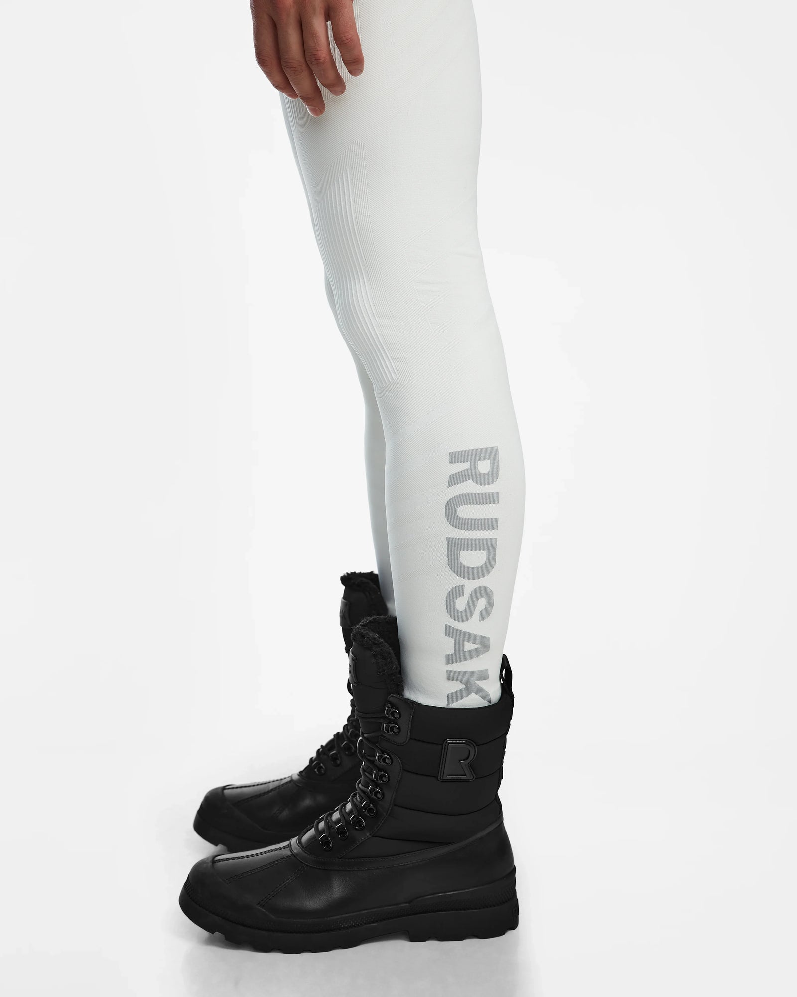 A person shown from mid-thigh down wears Rudsaks FRANC - 6524541 Iceland white stretch base layer leggings with vertical Rudsak branding and black lace-up winter boots, set against a white background.