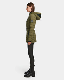 A woman in profile wears the Rudsak FRANCINE - 8124722 Loden Green puffer jacket with black leggings and black ankle boots, standing against a plain white background.