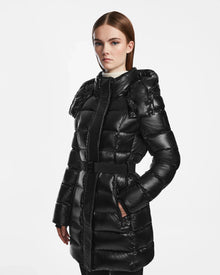 A woman with straight brown hair wears the Rudsak FRIDA S - L124526 Black, a shiny black quilted down jacket with a belted waist and removable hood, standing against a plain white background and looking at the camera.
