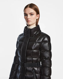 A person with straight brown hair wears the Rudsak FRIDA S - L124526 Black womens shiny mid-length down jacket with a high collar and belt, standing against a plain white background.