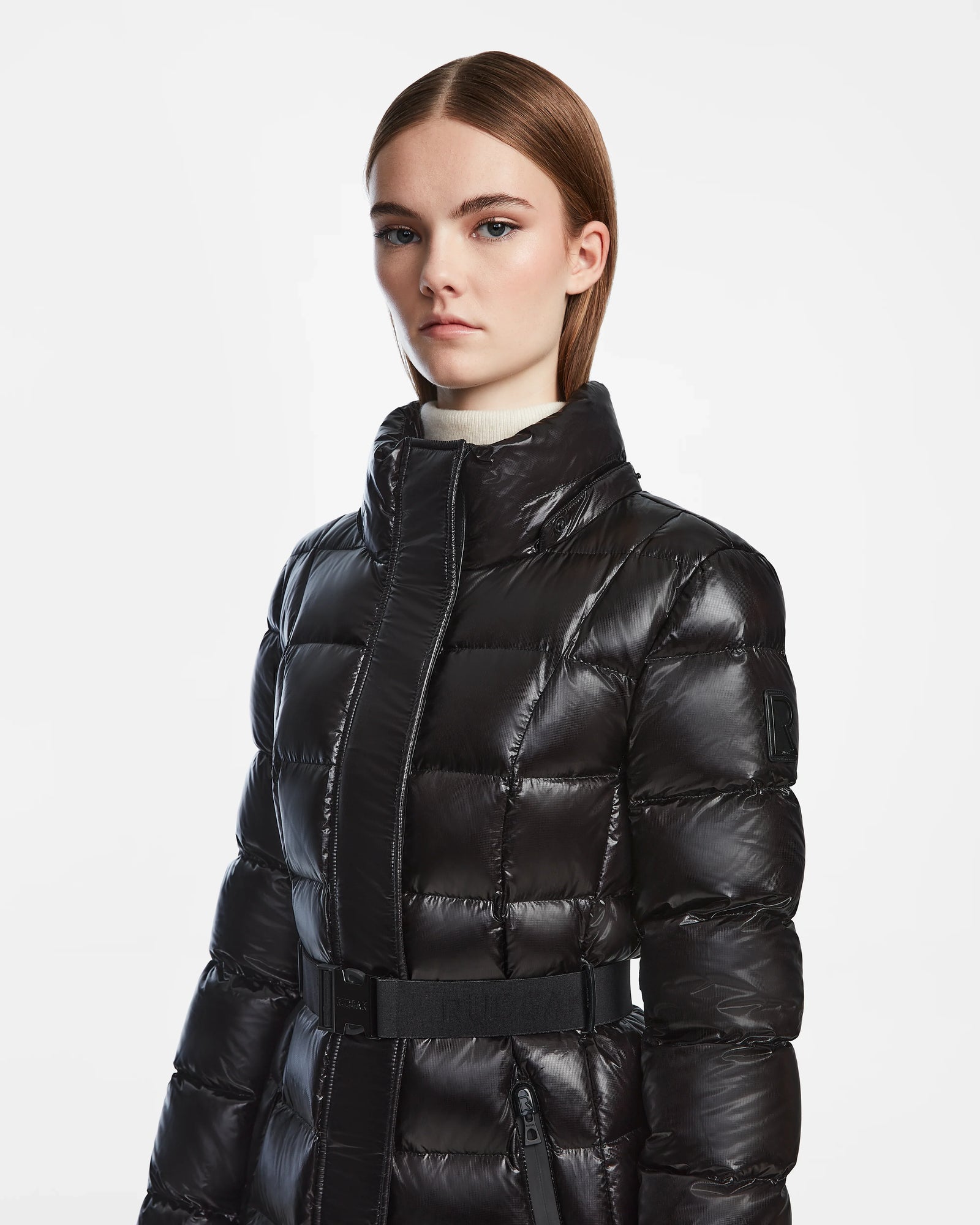 A person with straight brown hair wears the Rudsak FRIDA S - L124526 Black womens shiny mid-length down jacket with a high collar and belt, standing against a plain white background.