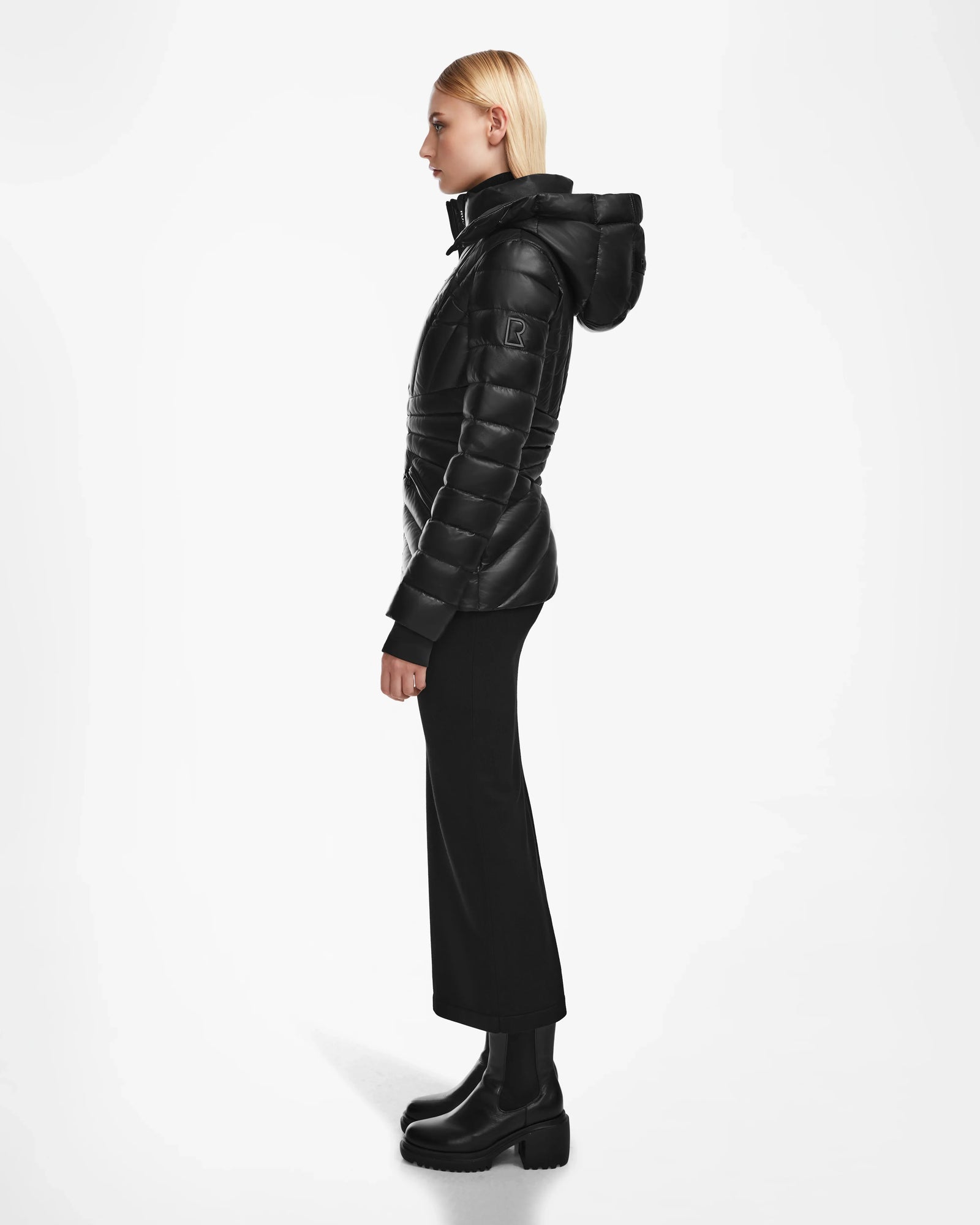 A person in profile wears the Rudsak HANNA X - 8123558 Black luxury down jacket with a hood, paired with black wide-leg pants and black platform boots, against a plain white background.