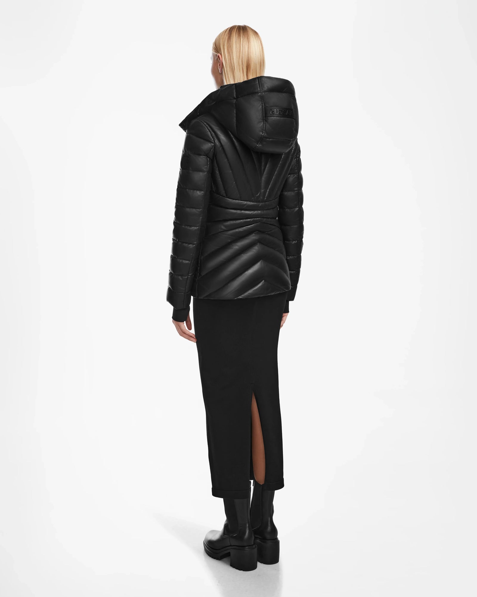 A person with blonde hair, wearing a black Rudsak HANNA X - 8123558 down jacket, a long black skirt with a back slit, and black boots, stands facing away against a white background.