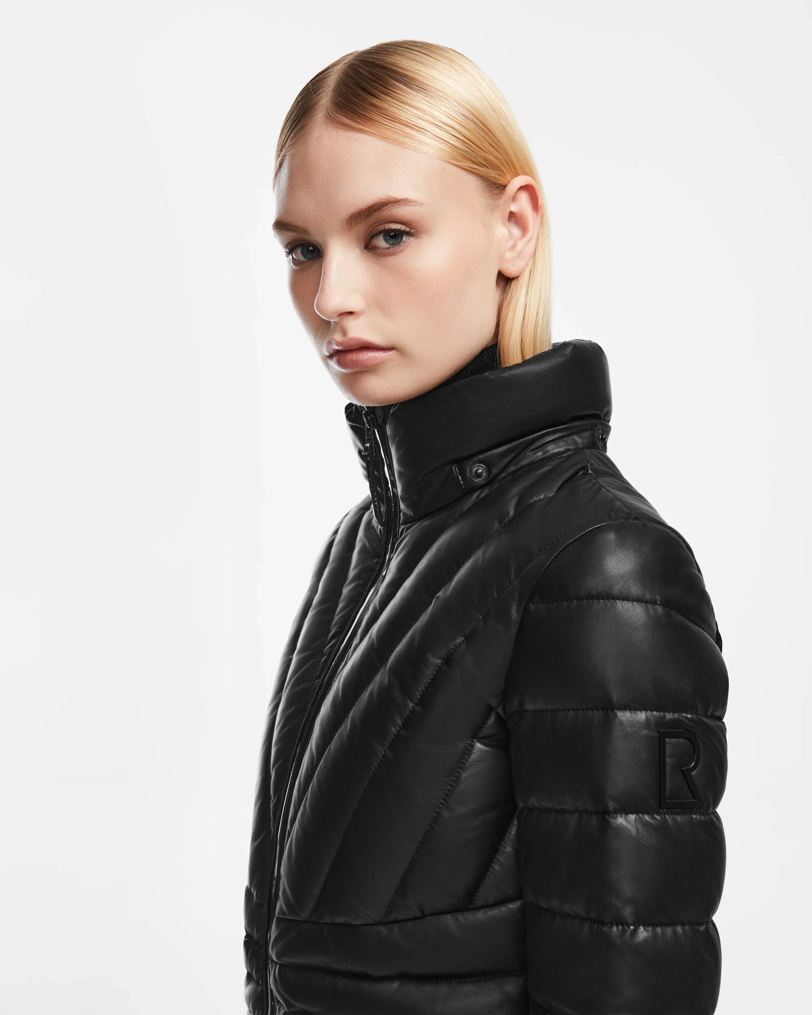 A woman with straight blonde hair models the Rudsak HANNA X - 8123558 Black women’s leather down jacket, showcasing its black quilted high-collar design and R logo on the sleeve, against a plain white background.