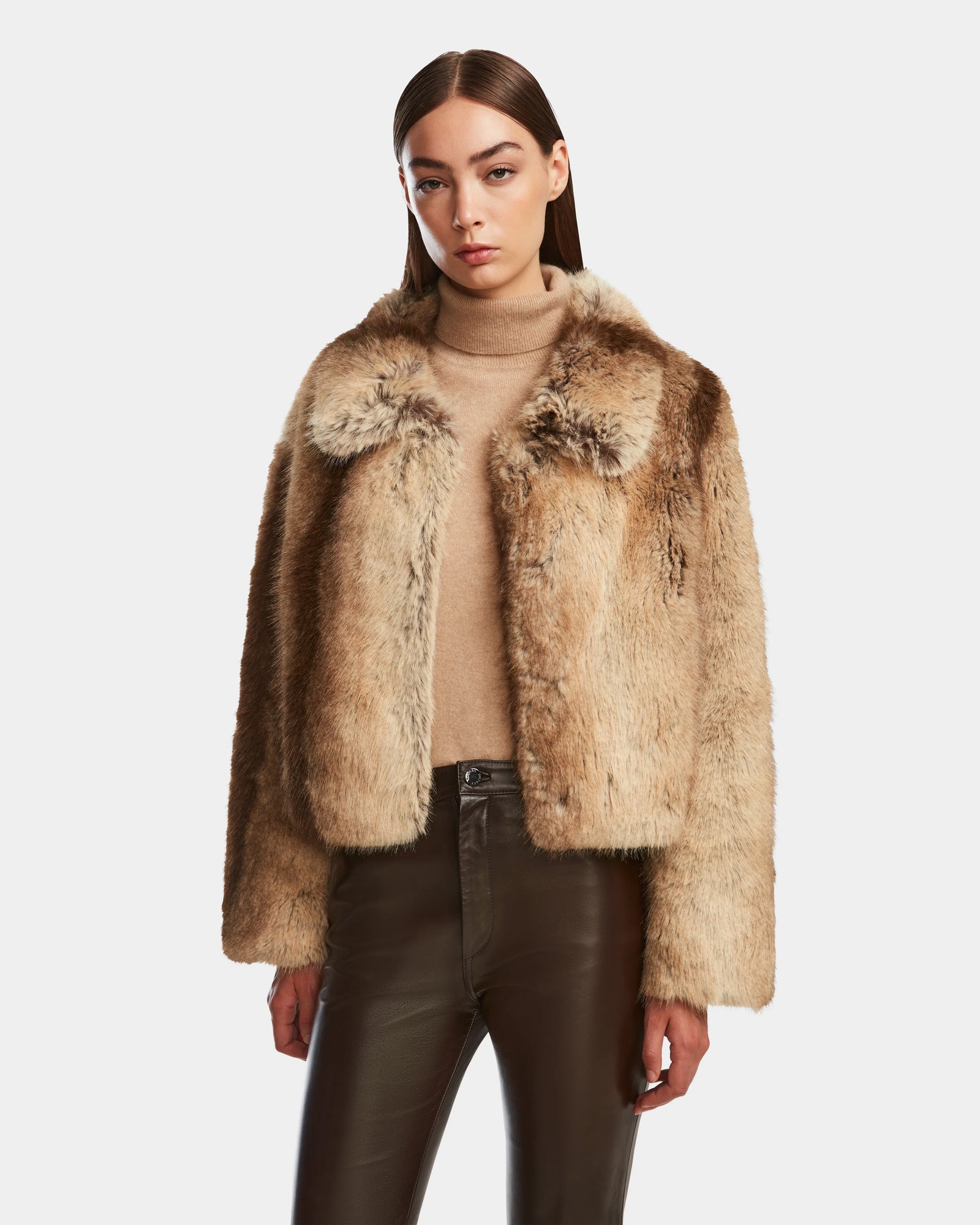 A woman with straight brown hair models the Rudsak JOSILYN - 8124792 Mocha women’s faux fur jacket over a beige turtleneck and dark leather pants, offering a chic winter look against a plain white background.