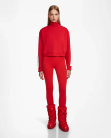 A woman wears the Rudsak KELLIE - 8524615 Flame Red mock neck top and matching leggings against a white background, styled with shiny red winter boots. The relaxed fit top is crafted from soft scuba fabric; her hair is straight and loose.