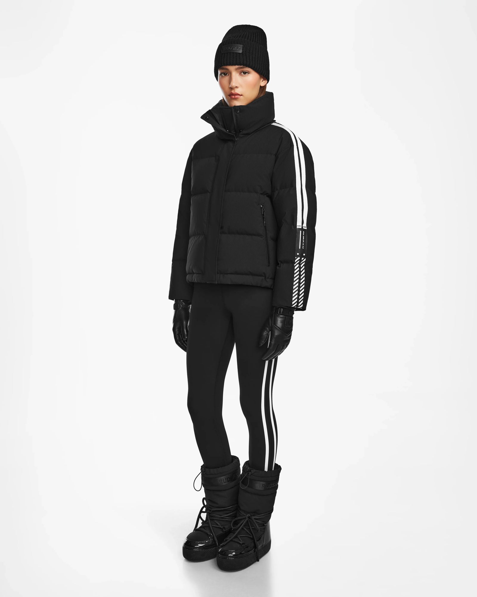 A person stands against a plain white background wearing the Rudsak KIMIA - 8124716 Black women's ski jacket with white sleeve stripes, paired with a black beanie, black gloves, black leggings with white side stripes, and black winter boots.