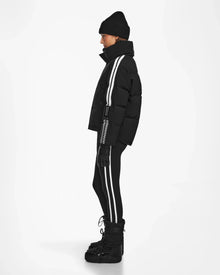 A person in profile wears a black beanie, gloves, winter boots, and the Rudsak KIMIA - 8124716 Black down ski jacket with bold white side stripes over matching leggings, providing stylish warmth against a plain white backdrop.