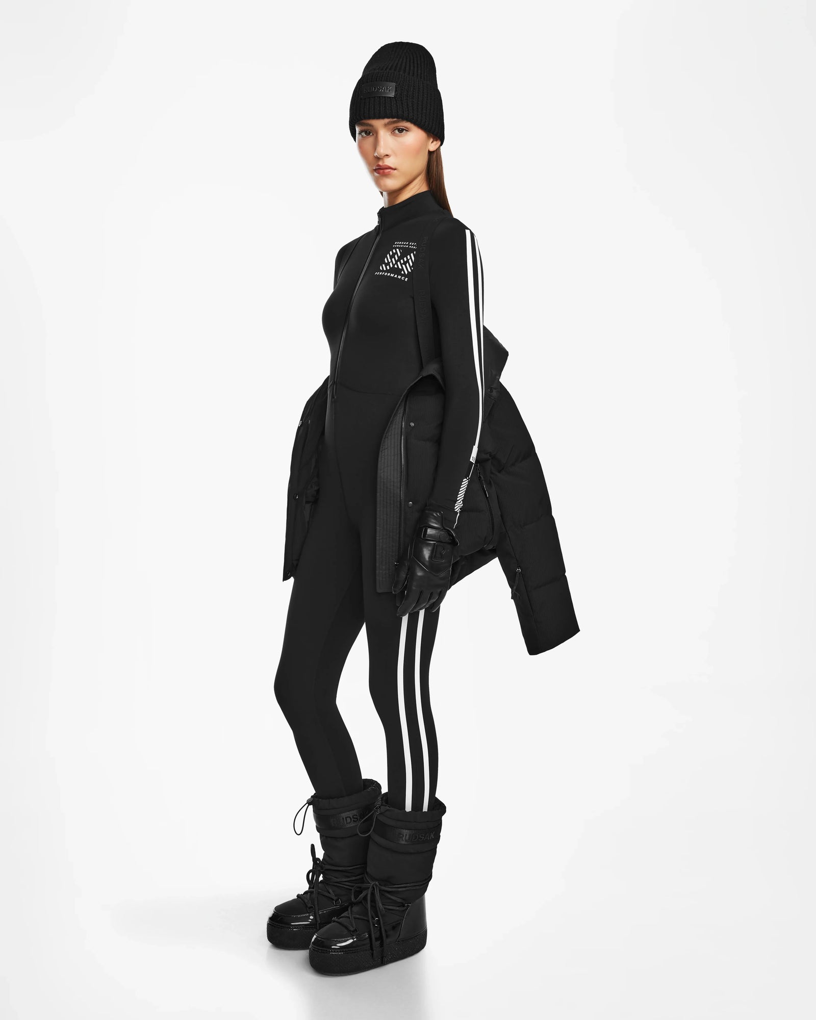 A woman in a black beanie, black fitted jumpsuit with white stripes, Rudsak KIMIA - 8124716 Black ski jacket, black gloves, and black snow boots stands against a plain white background.