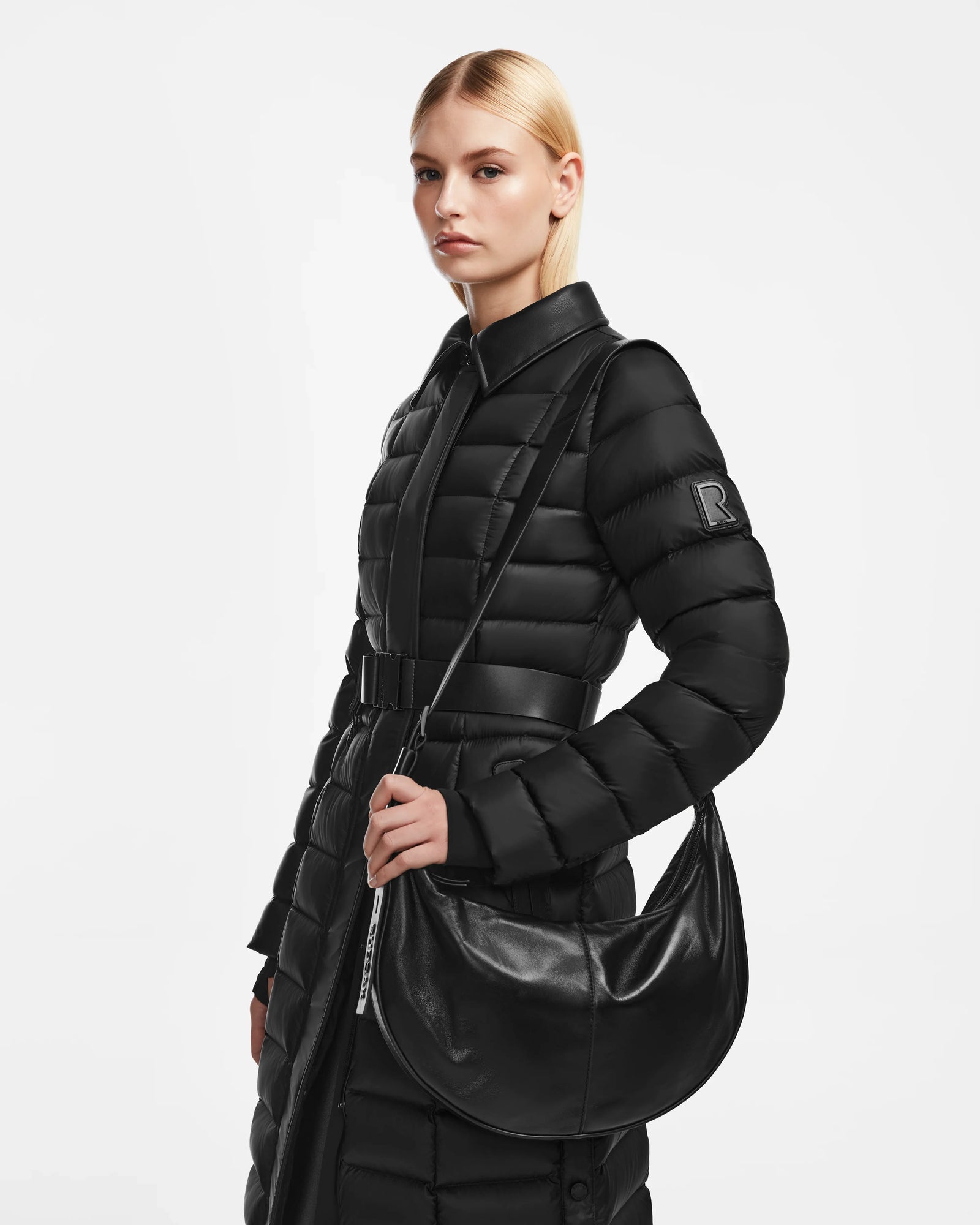 A woman with straight blonde hair, tucked behind her ears, models the Rudsak LANISSA - 8324505 Black puffer coat with belt and a black leather hobo bag, standing against a plain white background.