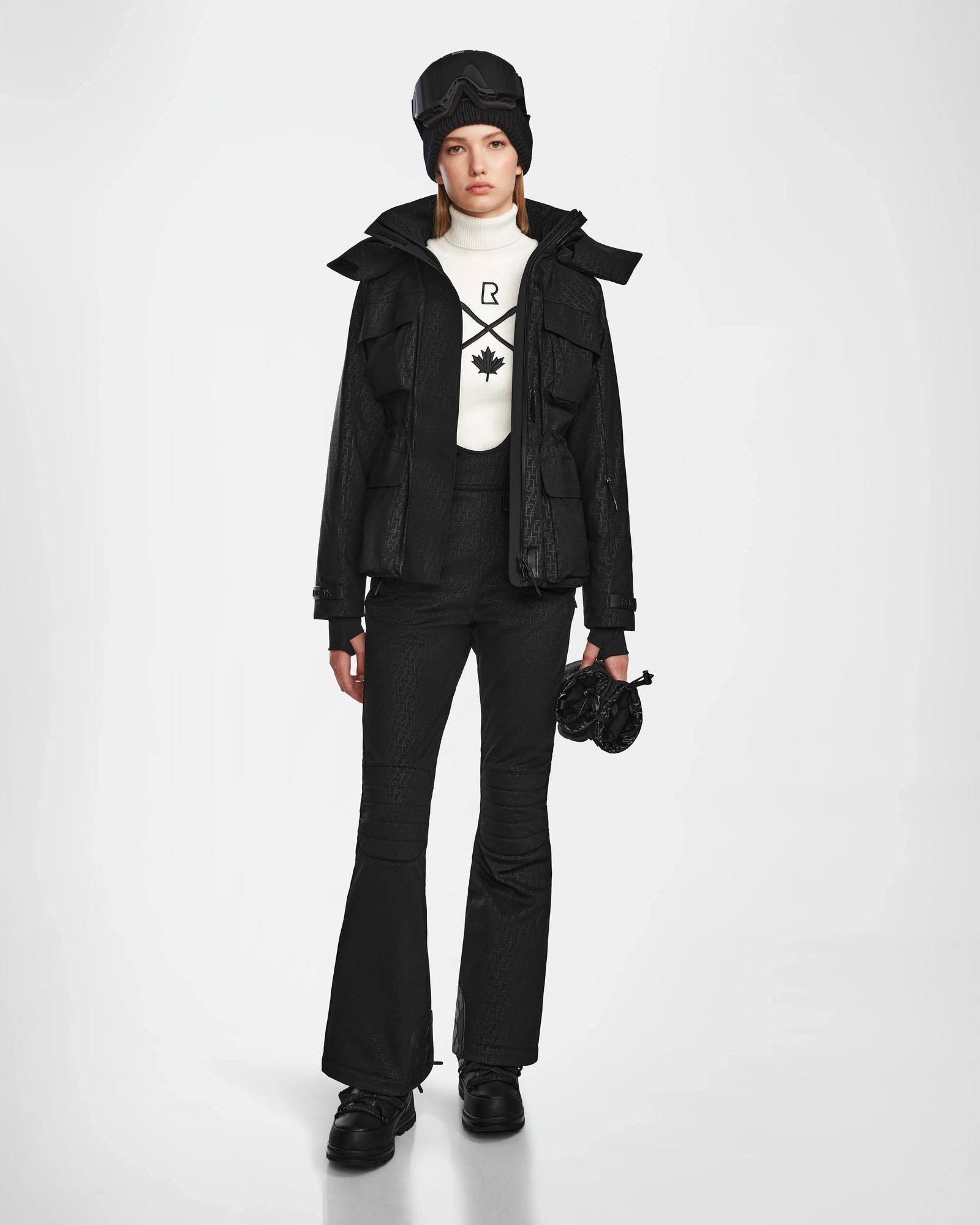 A person wears the Rudsak LUX MO - 8124570 Monogram Print Black womens ski jacket with black flared pants, boots, a hat, ski goggles, and a white turtleneck sweater featuring a black maple leaf design against a white background.