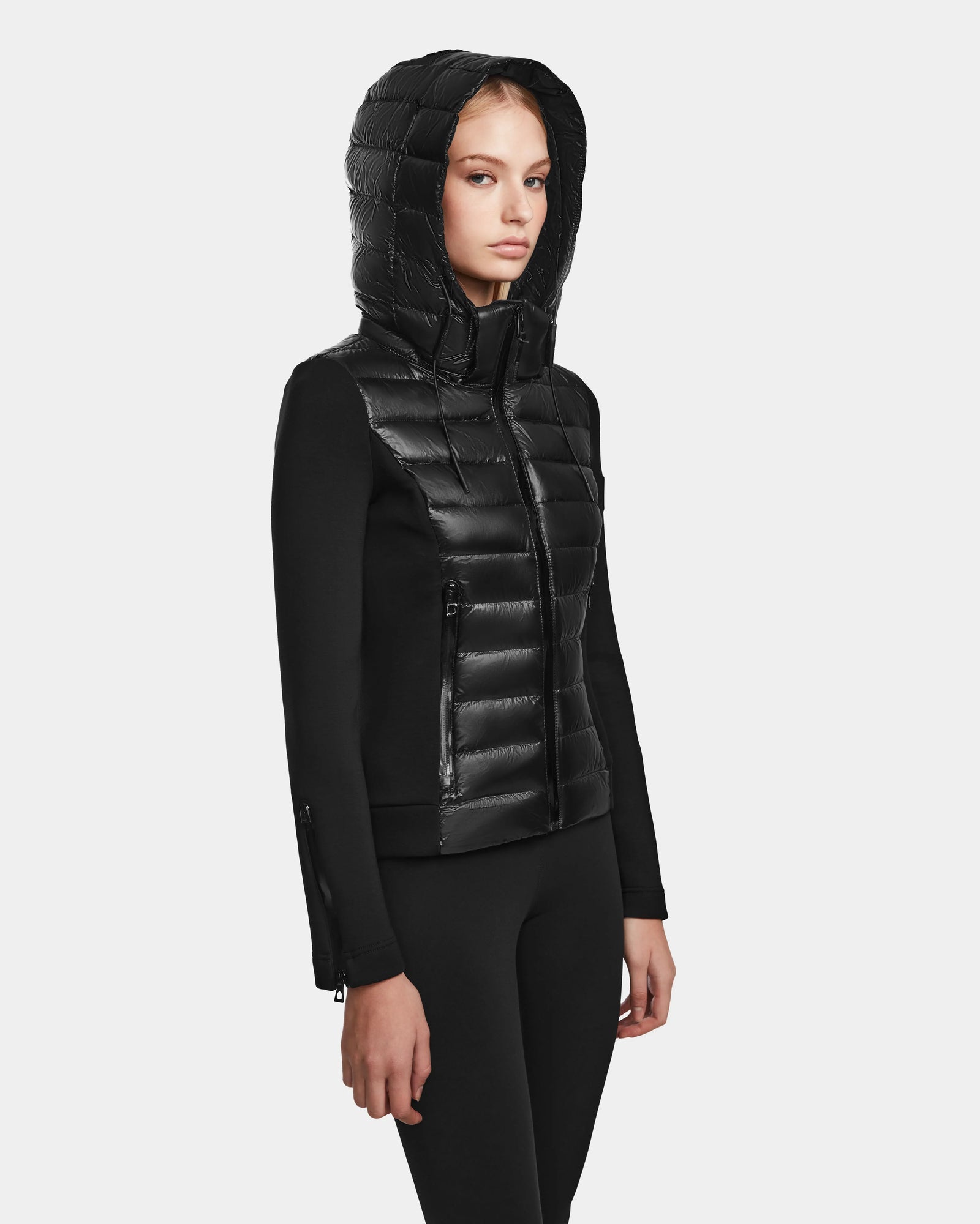 A woman wearing the RUDSAK MABS - 8122020 BLACK fitted womens puffer jacket with shiny quilted panels and matching black pants stands against a plain light gray background.