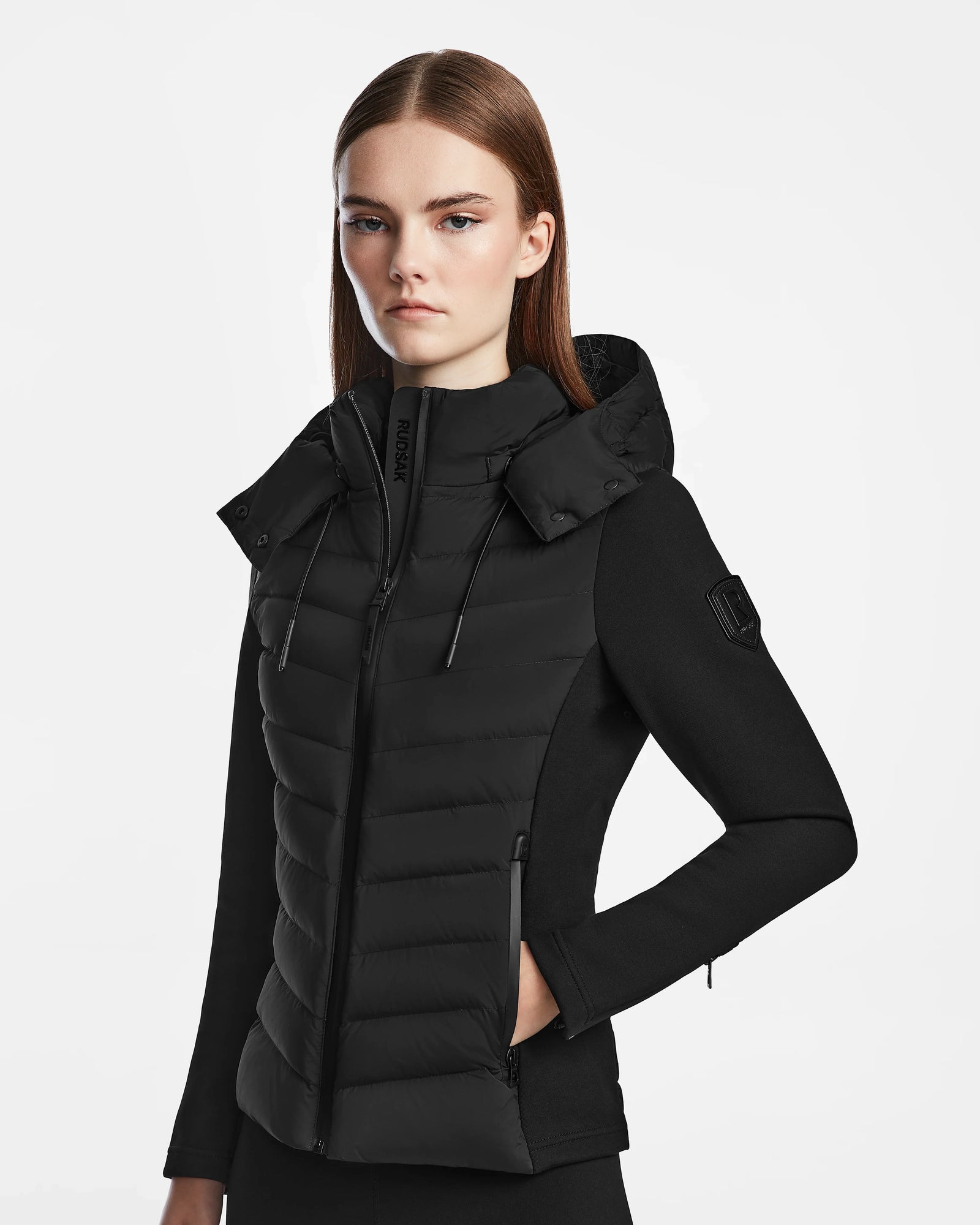 A woman with straight brown hair wears the Rudsak MALYA M - L123547 Black fitted down quilted jacket, featuring a high collar and hood, standing against a plain white background and looking directly at the camera with a neutral expression.
