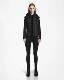 A woman faces forward in a Rudsak MALYA M - L123547 Black down quilted jacket with hood, black long-sleeve top, leggings, and ankle boots, set against a plain white background.