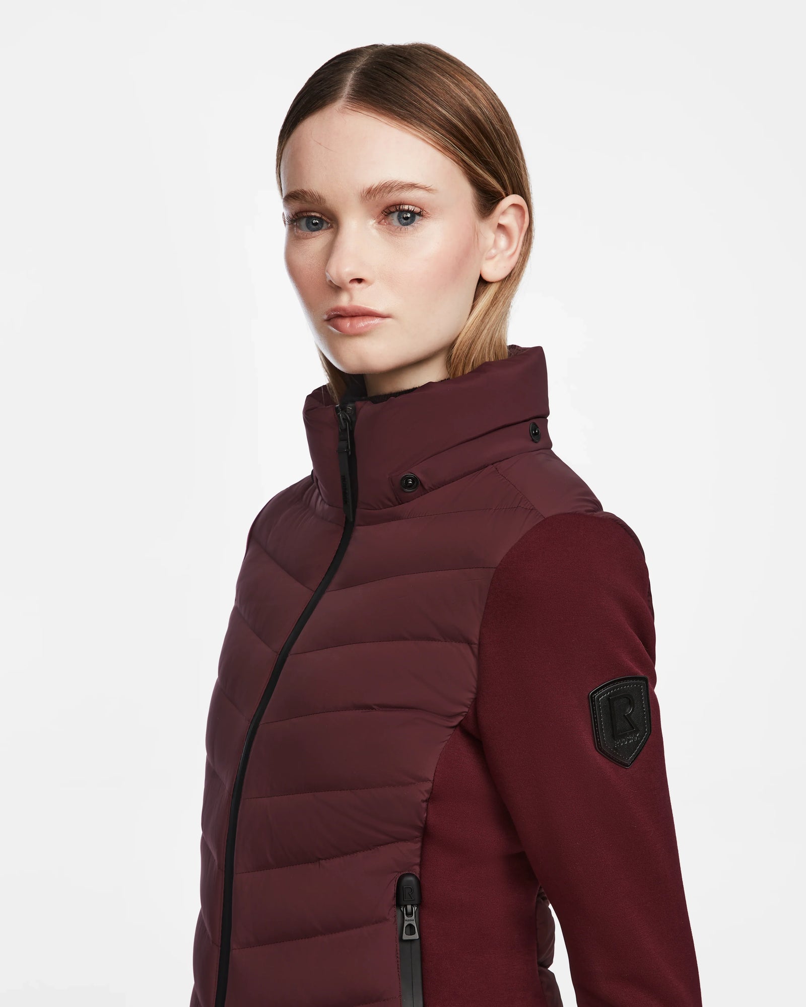 A woman with straight, light brown hair wears the Rudsak MALYA M - L123547 down quilted jacket in wine, featuring a high collar, zippered pockets, and a black sleeve patch. She stands against a white background, facing slightly left.