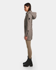 A woman in profile wears the Rudsak MERINA M - 8124030 Cement, a taupe quilted down jacket with a hood, paired with beige leggings and black lace-up boots, standing against a plain white background.