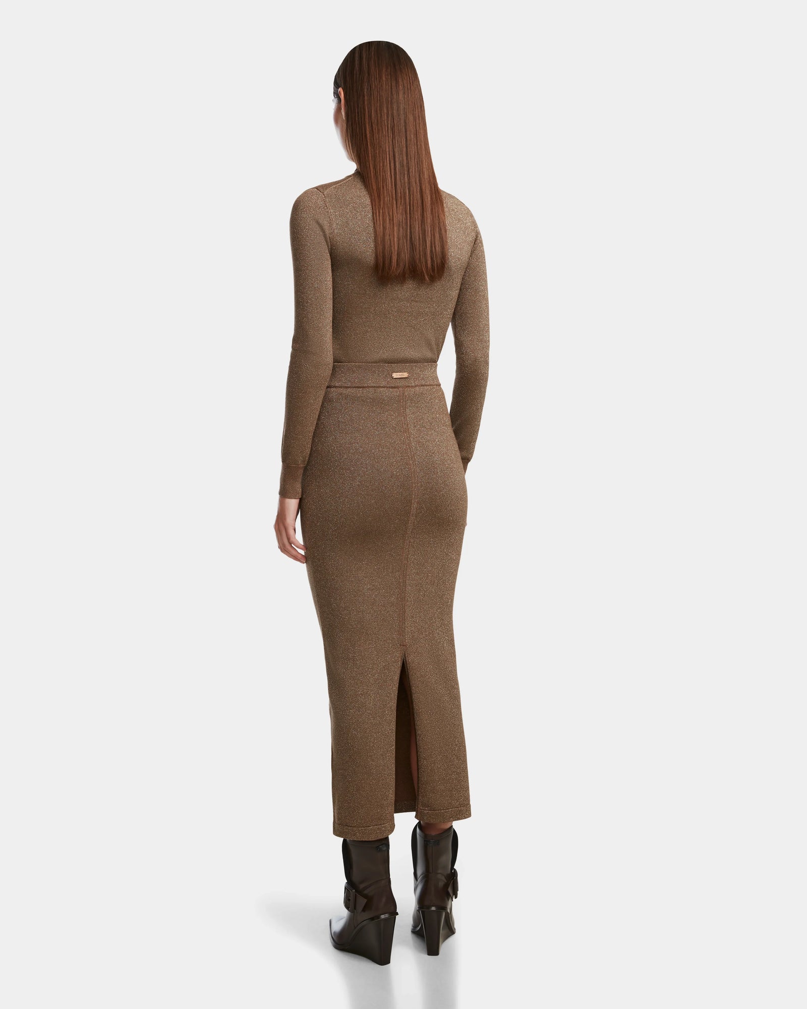 A woman with long straight brown hair wears the Rudsak MONICA - 8524516 Antique Bronze body-hugging, long-sleeved knit dress with a back slit and dark brown heeled boots, standing away from the camera to highlight its sleek silhouette.