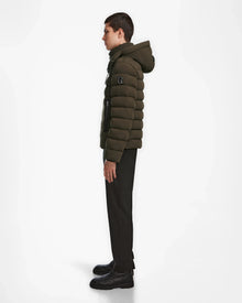 A young man stands in profile against a white background, wearing the Rudsak NAVAN - 6124551 Army mens light down puffer jacket with a hood, along with black pants and black shoes.