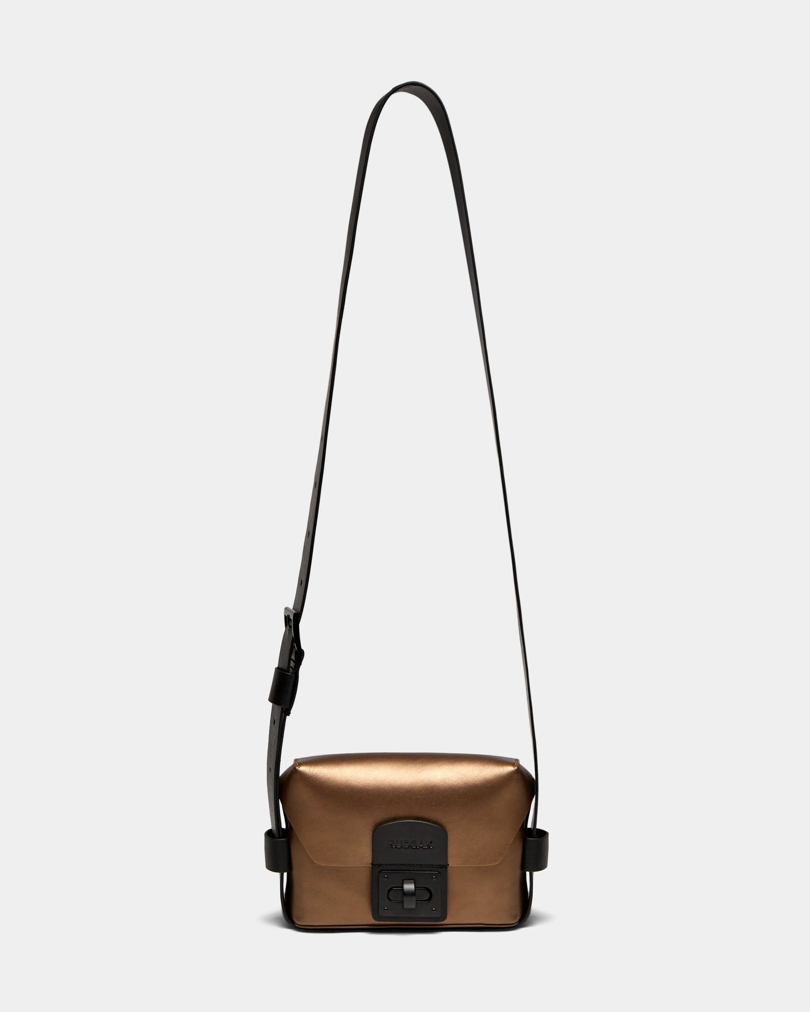 The Rudsak OFILIA - 8324504 is a compact, metallic antique bronze leather crossbody bag with a long adjustable black strap and front black buckle, shown on a plain light background.