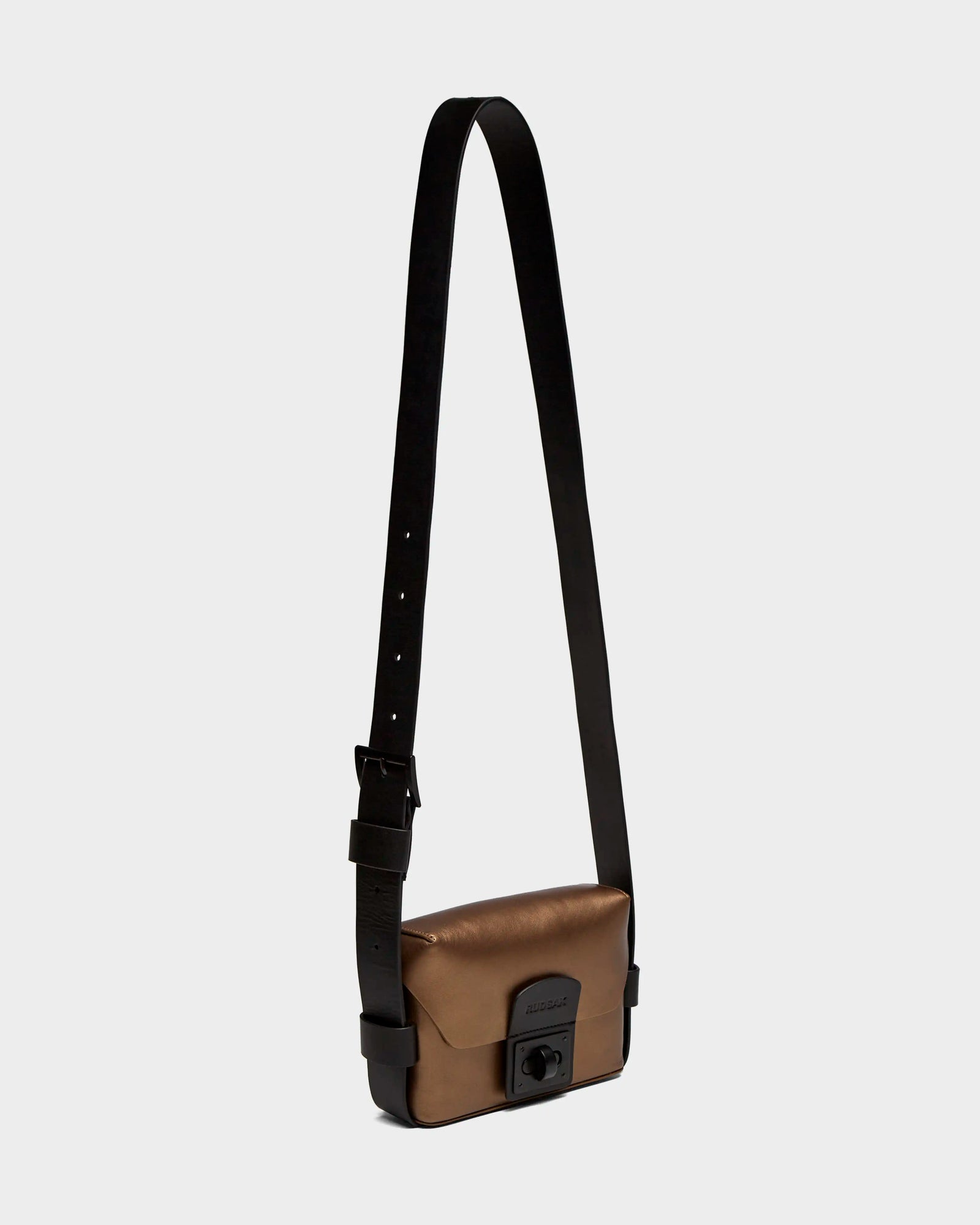 The OFILIA - 8324504 Antique Bronze by Rudsak is a small, rectangular metallic bronze leather crossbody bag with a wide adjustable black strap and a black front clasp, shown against a plain light gray background.