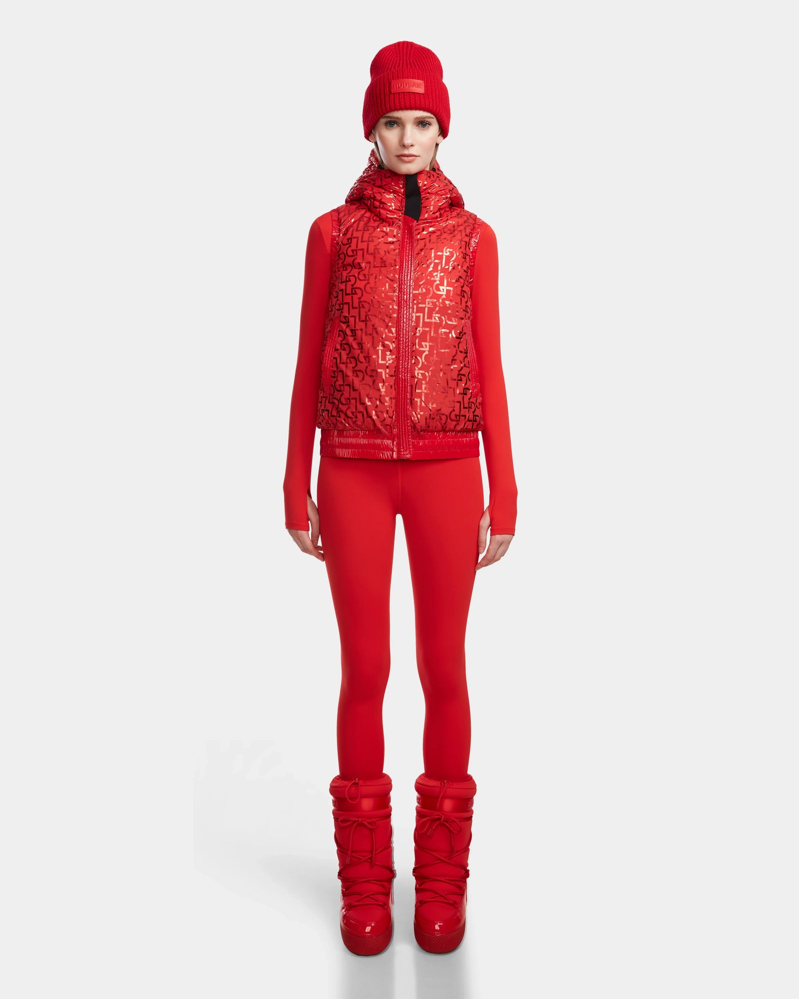A woman faces forward in a Rudsak RAQUEL M - 8124538 Flame Red high-performance reversible ski vest, matching leggings, beanie, and puffy boots, set against a plain white background.