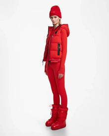 A woman poses against a white background in the Rudsak RAQUEL M - 8124538 Flame Red reversible ski vest, pairing it with a form-fitting outfit and matching red beanie and snow boots for a bold monochromatic look.