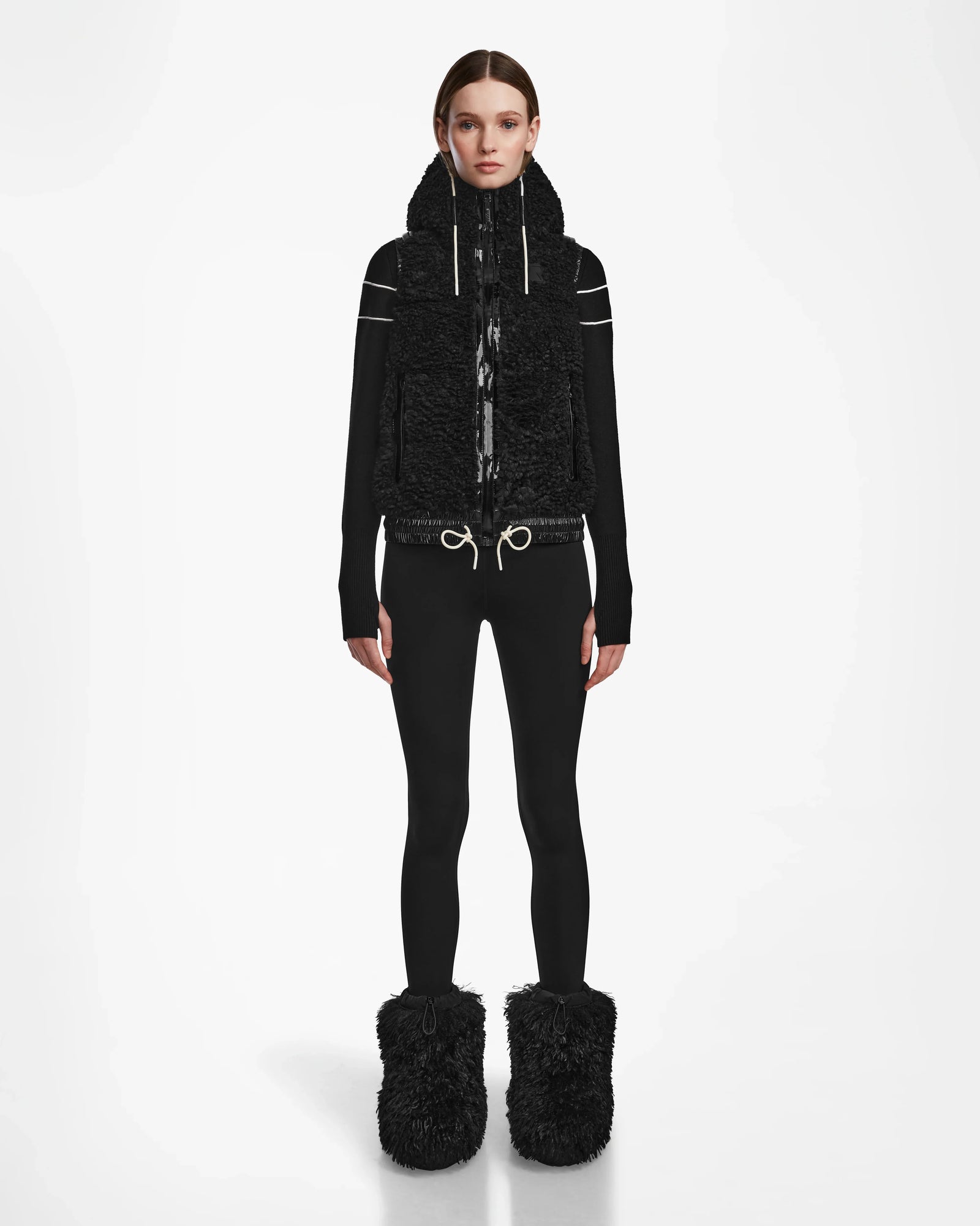 A person stands facing forward in the Rudsak RAQUEL S - 8124526 Black ski vest, black top, fitted pants, and large black fuzzy boots—ready for winter mountain adventures against a plain white background.