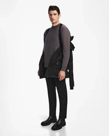 A man stands against a white background, wearing a gray sweater, black pants, black boots, and carrying the Rudsak RICKY - M123502 Black parka over one shoulder. He looks forward with a neutral expression.