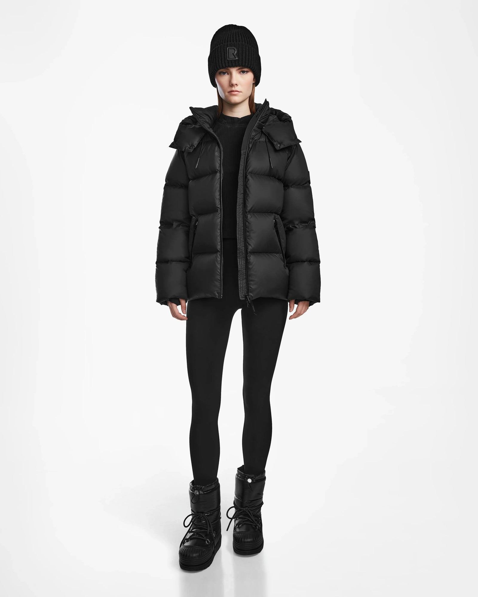 A person faces forward against a white background, wearing the Rudsak RITA M - L124537 Black weather-resistant puffer jacket, styled with a black beanie, black leggings, and black boots.