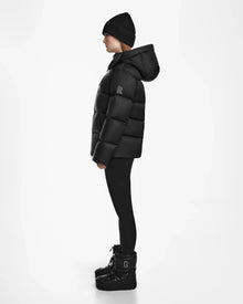 A person in profile stands against a white background, wearing the Rudsak RITA M - L124537 Black weather-resistant puffer jacket with a hood, paired with a black beanie, black leggings, and black winter boots.