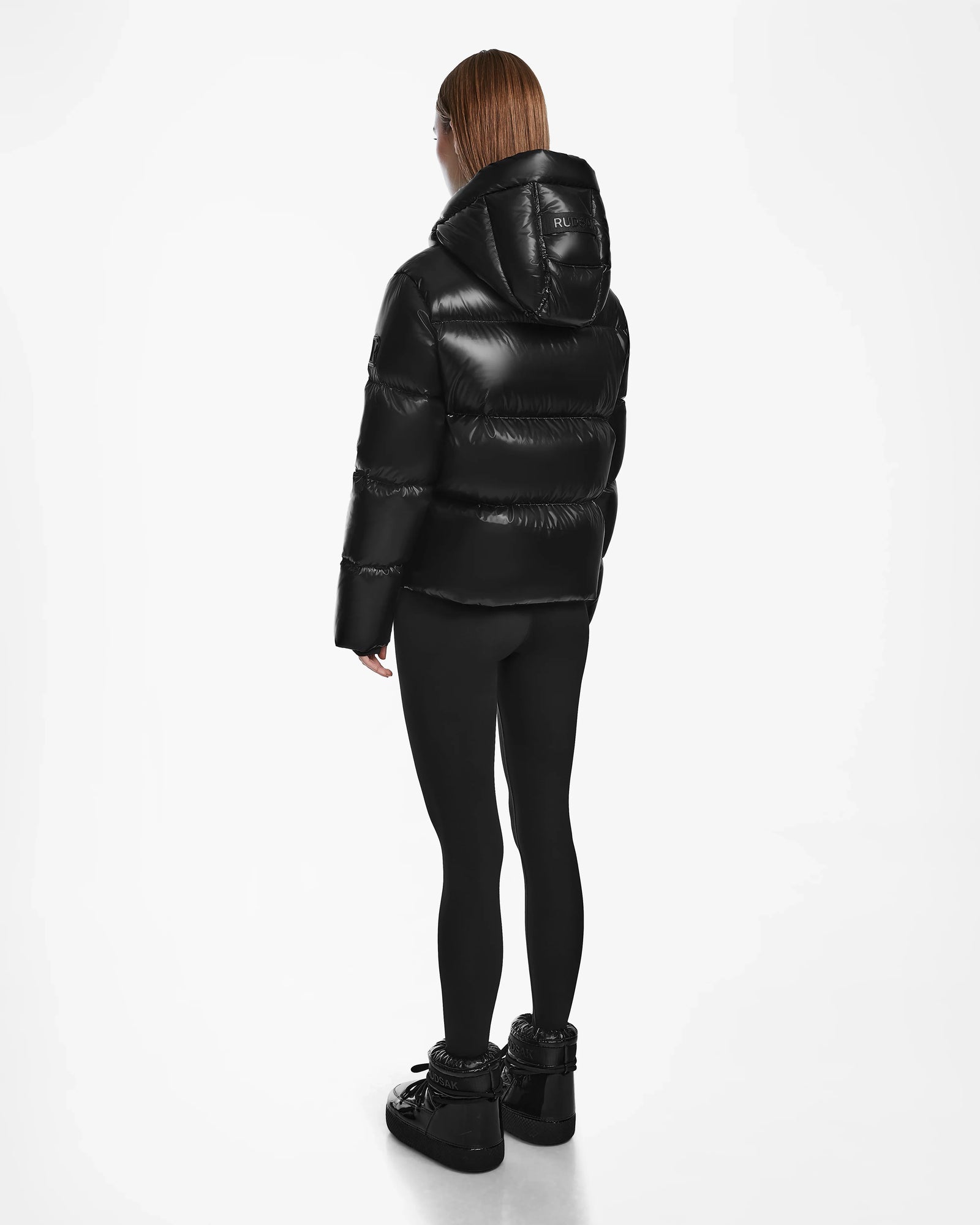 A person with straight, shoulder-length hair models the Rudsak RITA S - L124520 Black weather-resistant puffer jacket, paired with black leggings and sneakers, standing and facing away on a plain white background.