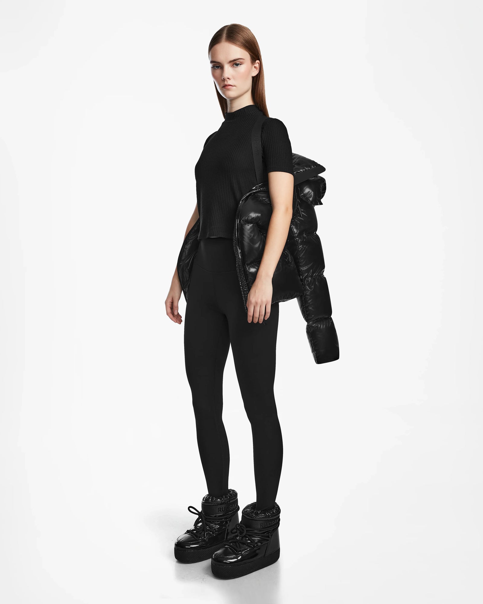 A woman with long straight hair and a neutral expression stands against a white background, wearing the Rudsak RITA S - L124520 Black puffer jacket draped over one shoulder, paired with a black top, leggings, and puffer boots.