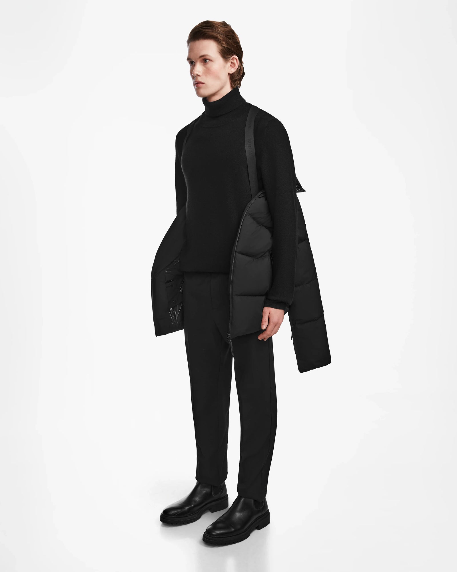 A person stands facing left against a white background, wearing a black turtleneck, pants, shoes, and Rudsak’s THEO - M125531 Black men’s down puffer jacket draped over their shoulders.