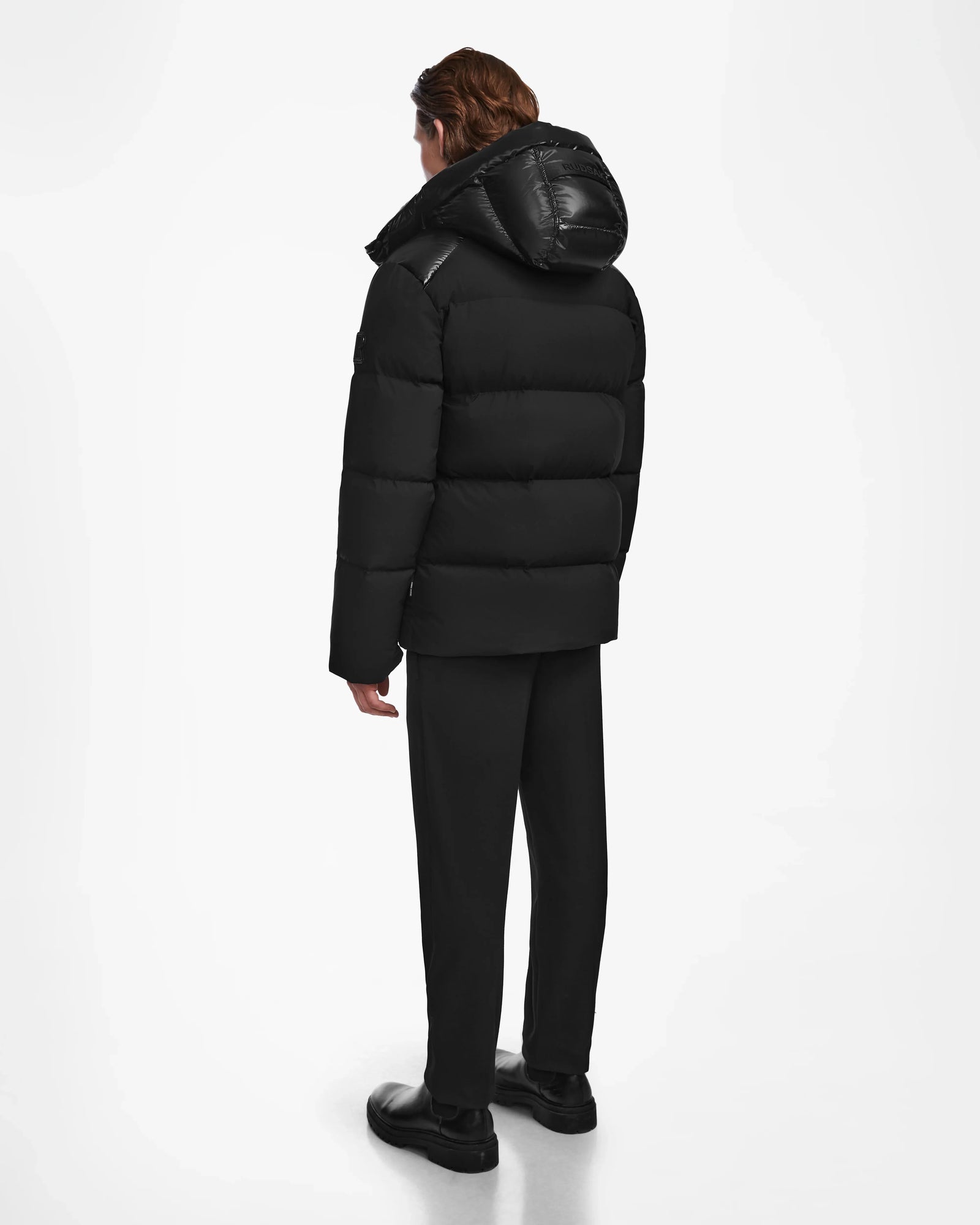 A person stands facing away, wearing the Rudsak THEO - M125531 Black men's down puffer hooded jacket, paired with black pants and shoes against a plain white background.