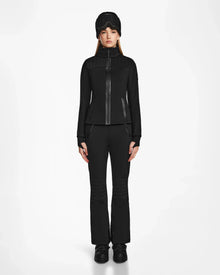A woman faces forward wearing the Rudsak ROSALIE - 8124784 Black fitted ski jacket with matching black pants, boots, and a black winter hat, set against a plain white background.