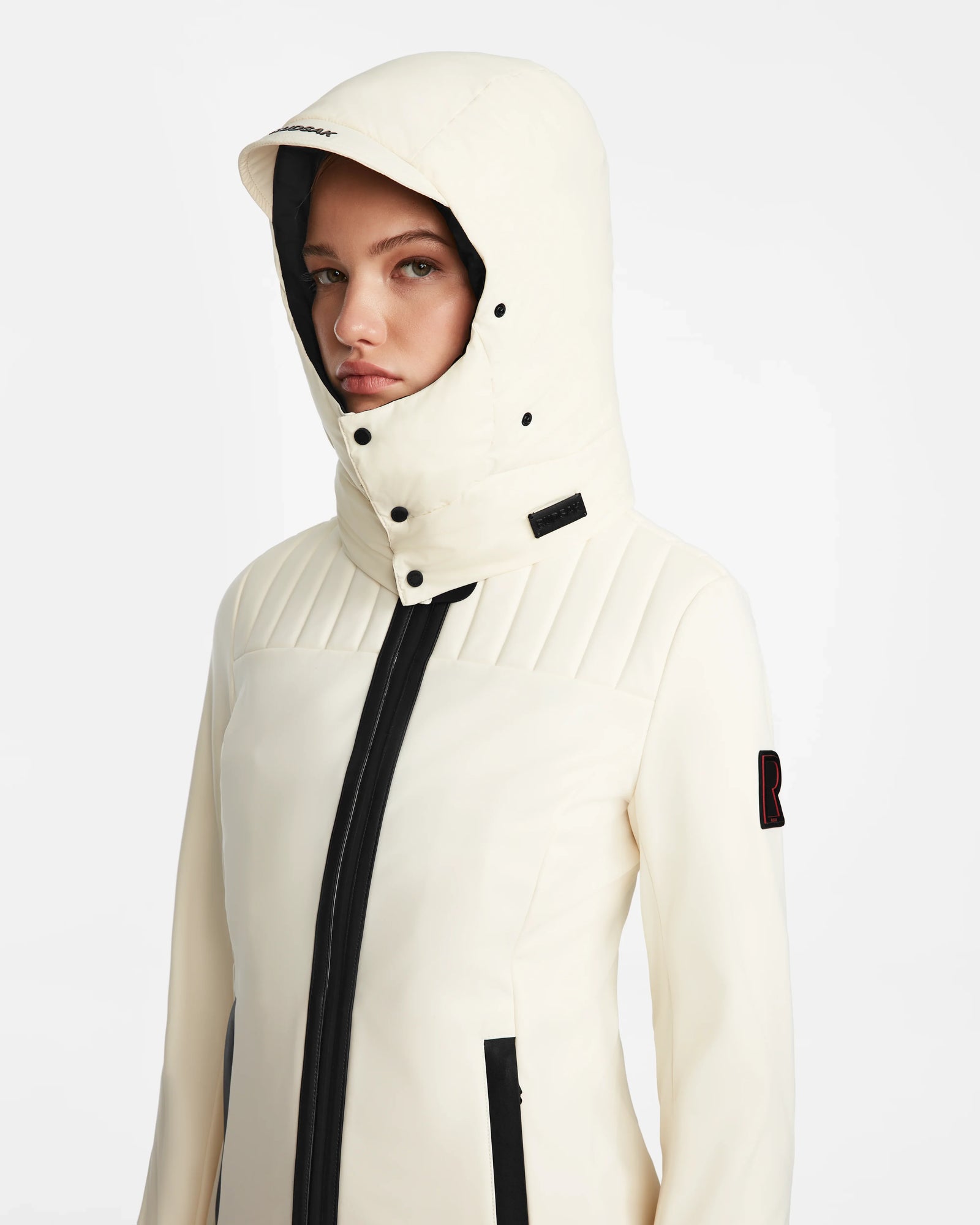 A woman wearing the Rudsak ROSALIE - 8124784 Iceland white fitted ski jacket with a high collar, black accents, and zipper, stands against a plain white background.