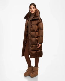 A woman wears the Rudsak SASH R - 8124543 Antique Bronze long down puffer coat with RDS-certified down, paired with a brown turtleneck, matching leggings, and tan boots, standing against a plain white background.
