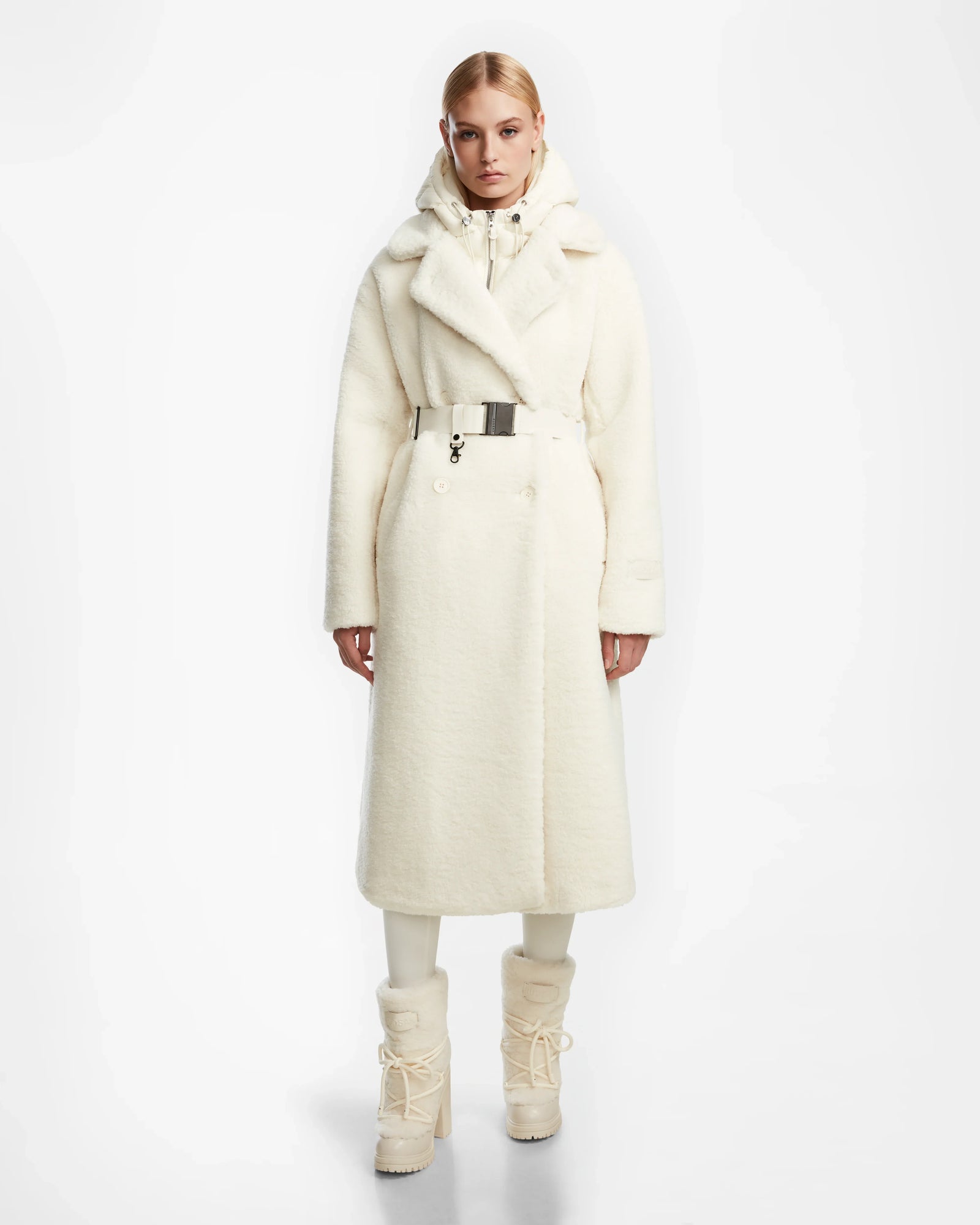 A woman faces forward in a long, white Rudsak TINA - 8123578 Iceland faux fur teddy trench coat with a wide collar and silver belt, styled with white pants and boots against a plain white background.