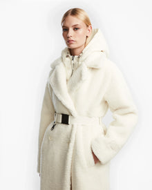 A woman with blonde hair models the Rudsak TINA - 8123578 Iceland: a white belted faux fur trench coat with a large collar and removable hooded bib. She stands against a plain white background, one hand in her pocket.