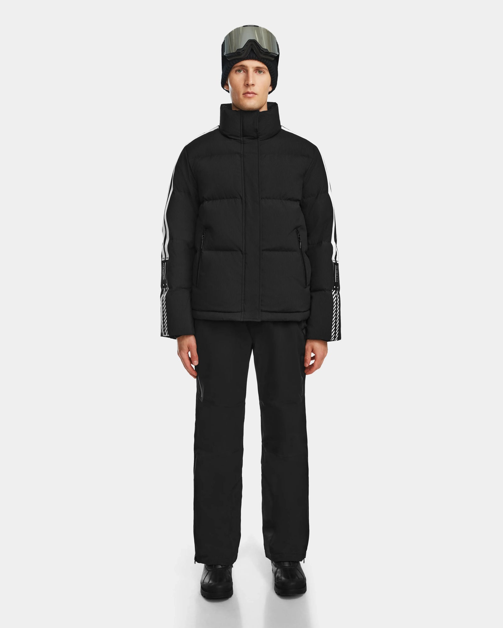 A person wearing the Rudsak USHER - 6124516 Black water-resistant ski jacket and pants, black boots, a black beanie, and ski goggles stands facing forward against a plain white background.