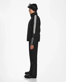 A man in profile wears the Rudsak USHER - 6124516 Black men’s down ski jacket with white sleeve stripes, black pants, boots, and a beanie with goggles on top. A plain white background emphasizes the sleek winter style.