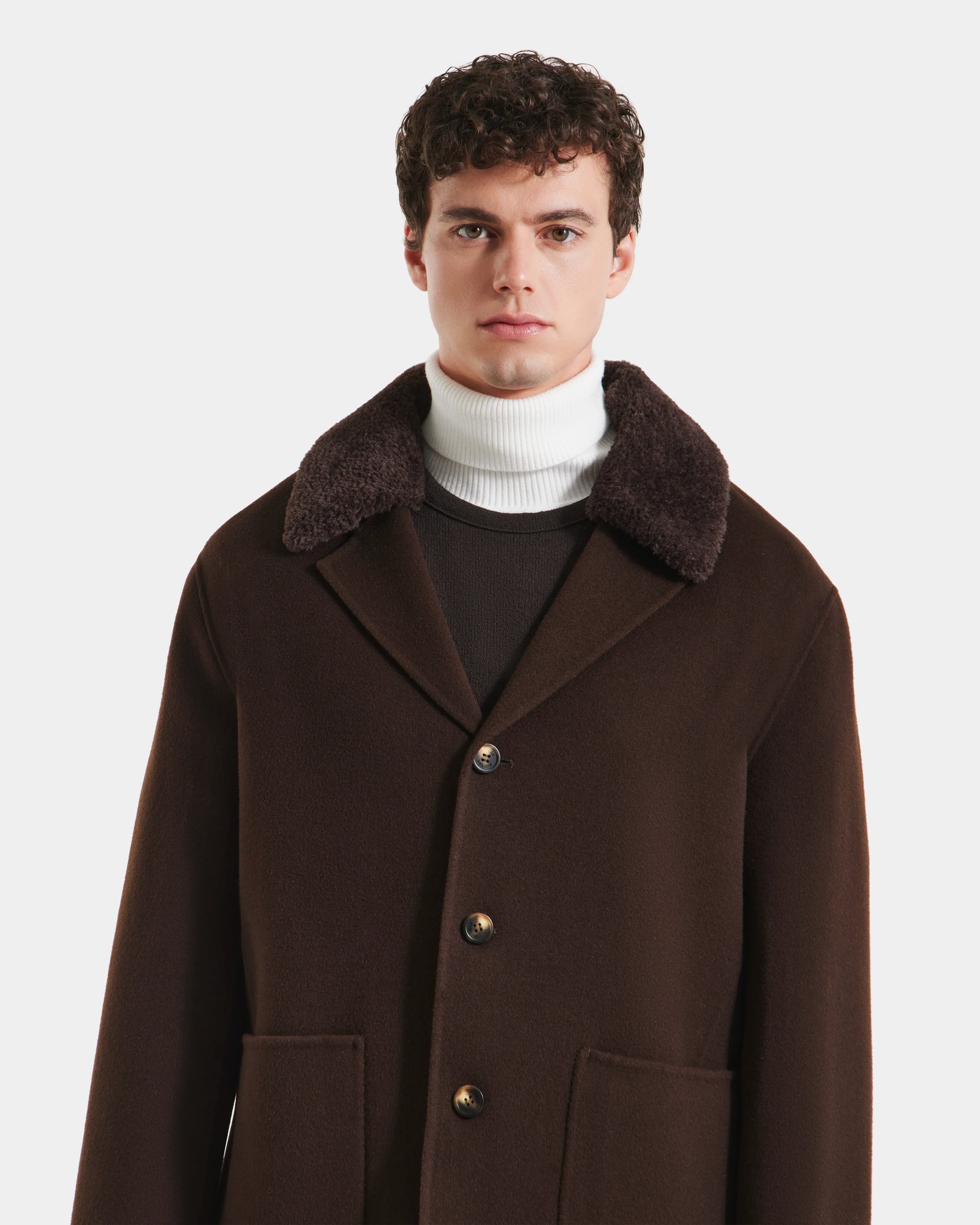 A man with short curly hair models the Rudsak AIDEN - 6125558 Espresso wool-blend coat, featuring a wide removable shearling collar, layered over a white turtleneck against a plain white background.