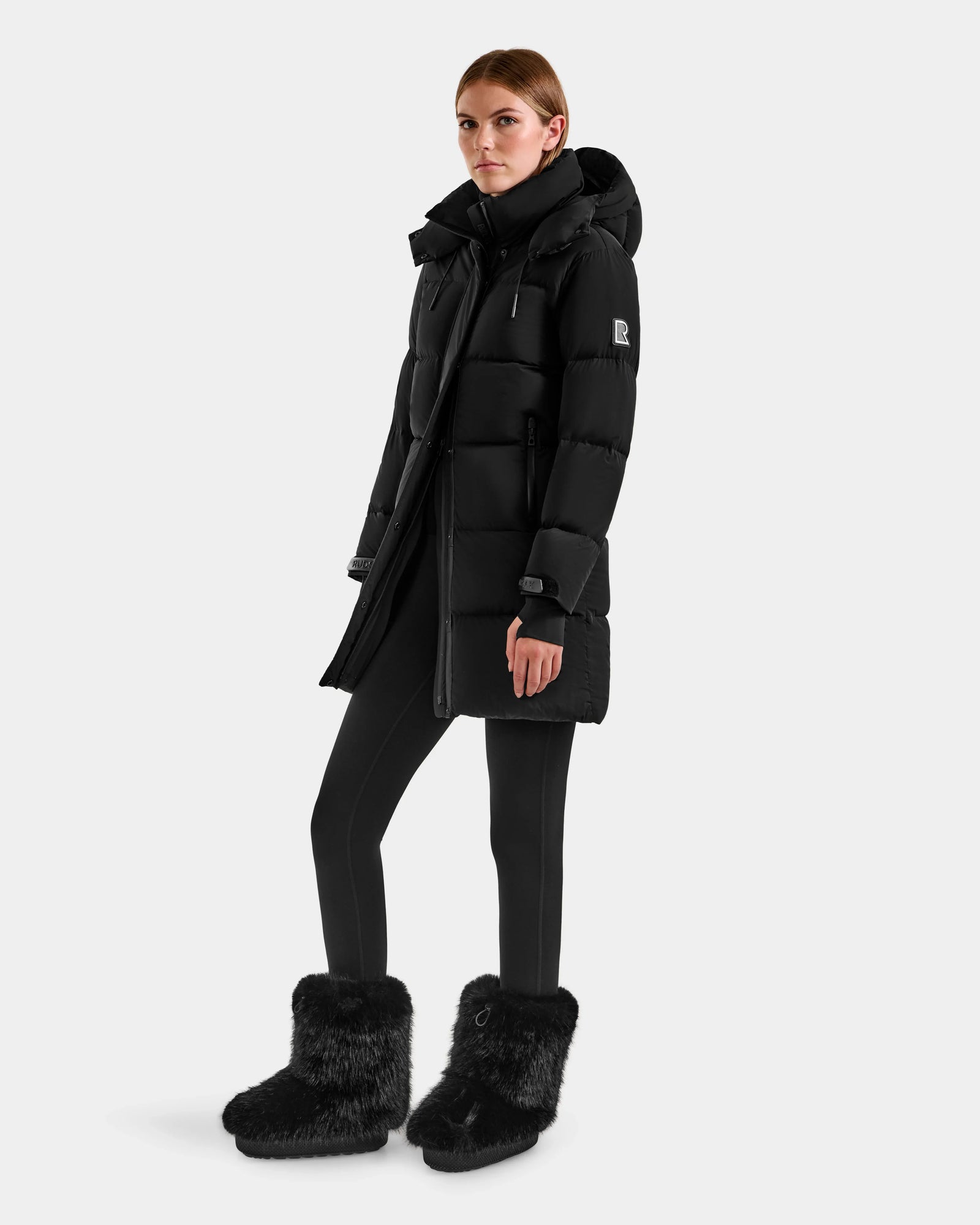 A woman with light brown hair pulled back and a neutral expression wears the Rudsak ALESSANDRA M - L125552 Black down puffer coat, paired with black leggings and black furry winter boots against a plain background.