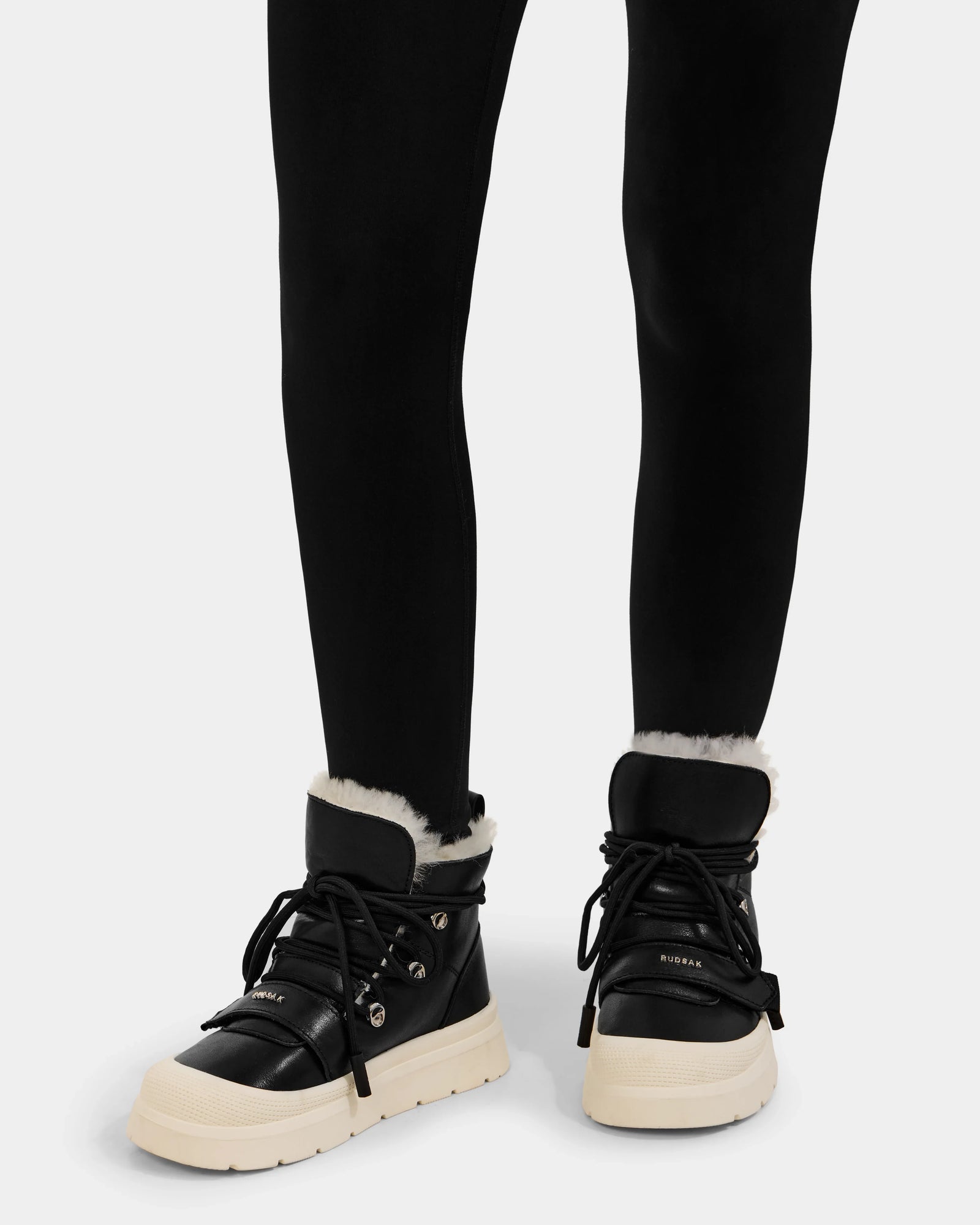 A person wearing Rudsak ALEXEY L - L225518 Black winter boots with white soles and visible wool lining, paired with black leggings, standing against a plain background.