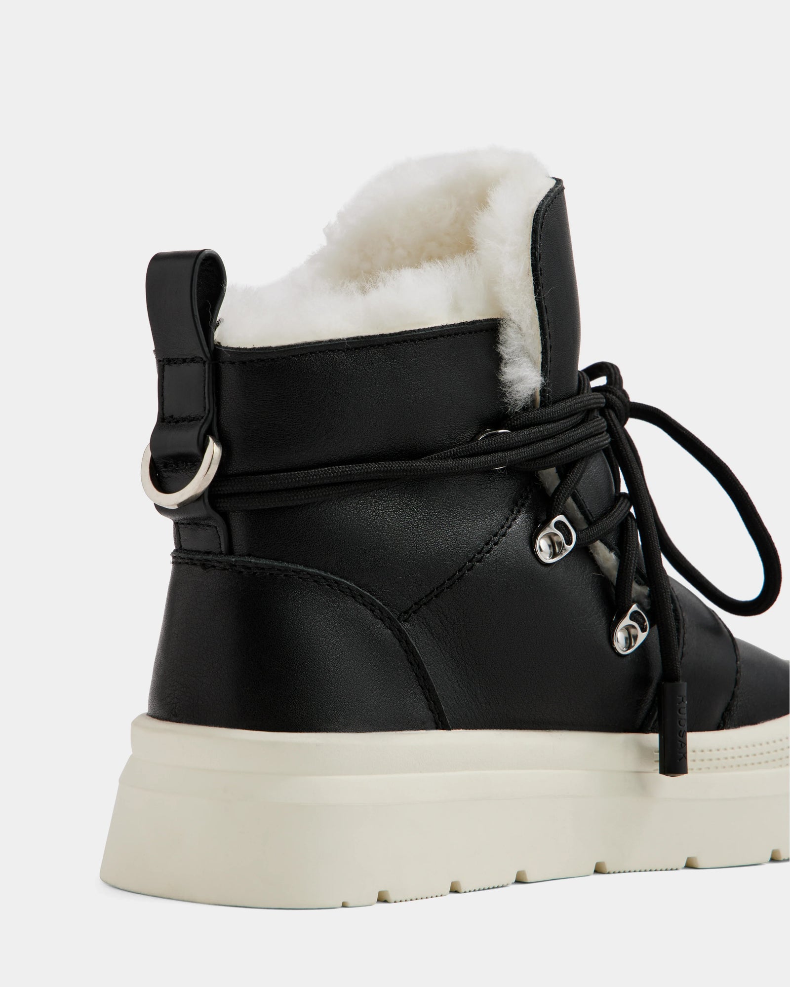 A close-up of the Rudsak ALEXEY L - L225518 Black women’s leather boot featuring black lace-up detail, plush white wool lining, and a thick white sole, shown from the back and side against a plain background.
