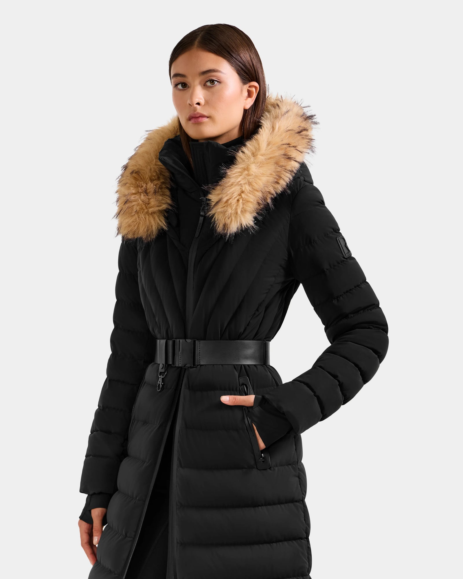 A woman models the Rudsak ALISA L - L125574 Black long quilted puffer coat with a fur-lined hood and vegan-leather belt, standing with one hand in her pocket against a plain light background.