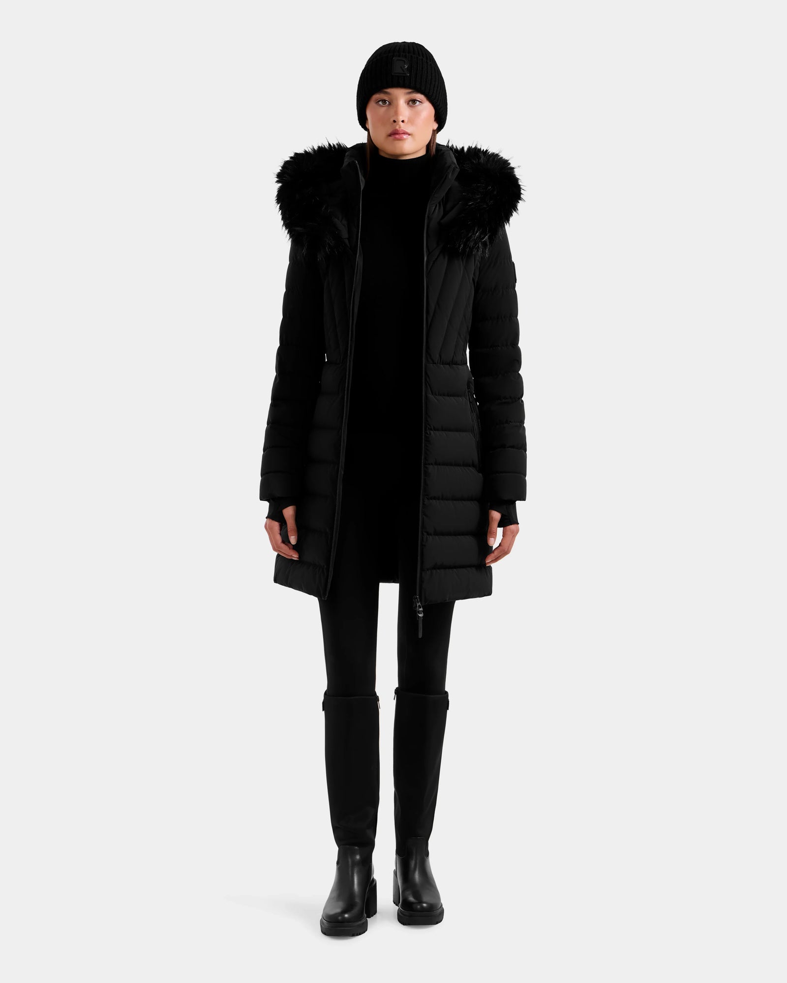 A person stands facing forward against a light background, wearing the Rudsak ALISA M - L125586 Black quilted down parka with a fur-trimmed hood, paired with a black beanie, pants, and boots.